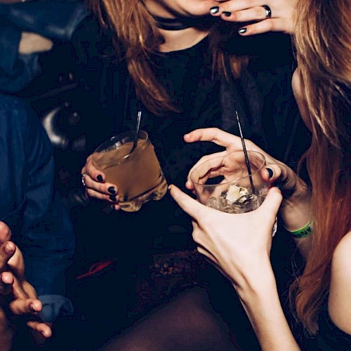 Two women at a party hold smoky cocktails with straws; hands show black rings and tattoos as they enjoy drinks.