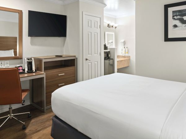 A modern hotel room with a desk, chair, flat-screen TV, dresser, bed, and a mini-fridge area near a doorway, all in neutral tones.