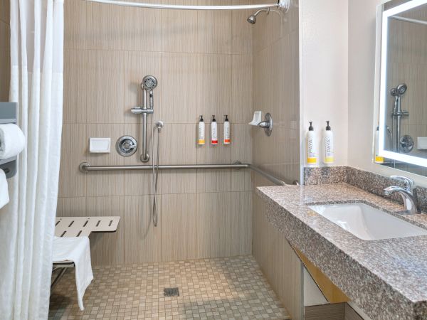 A modern hotel bathroom with a rainfall shower, soap dispensers, and a granite sink counter with chrome fixtures.