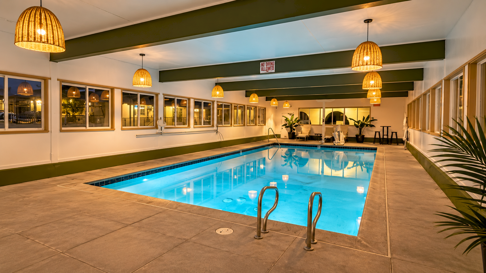An indoor swimming pool with blue water, golden pendant lights, lounge chairs, and a &ldquo;no diving allowed&rdquo; sign above.