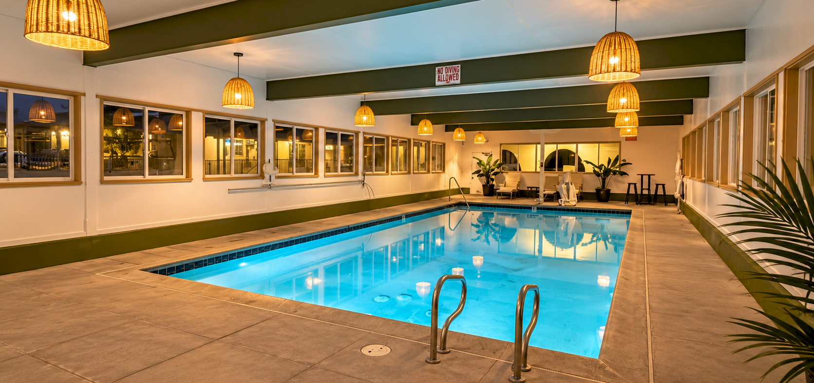 An indoor swimming pool with blue water, golden pendant lights, lounge chairs, and a &ldquo;no diving allowed&rdquo; sign above.