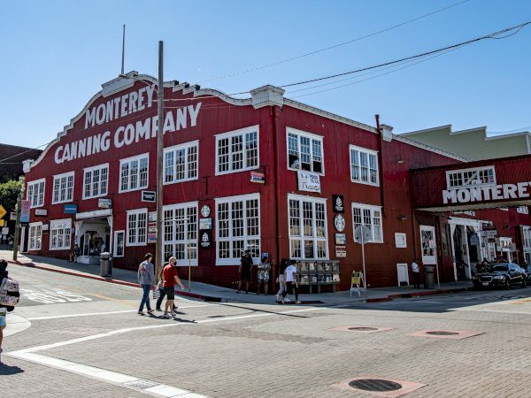 A red, two-story building labeled &ldquo;Monterey Canning Company&rdquo; sits on a street corner with people walking nearby under a clear sky.