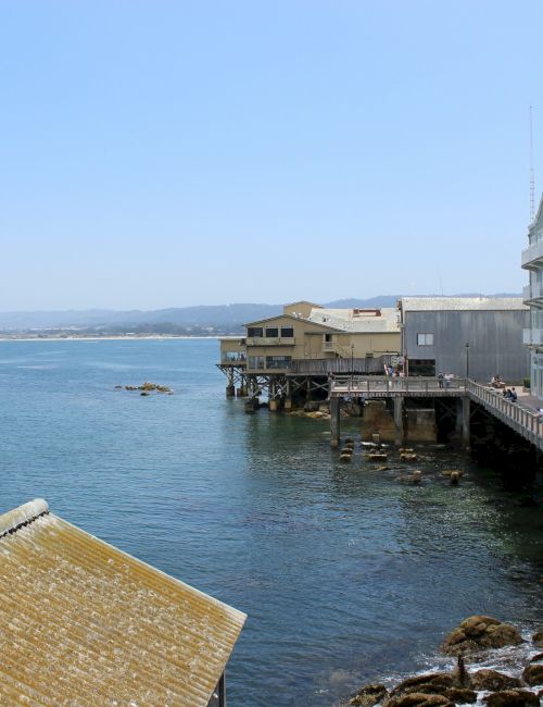 A seaside view from a deck overlooking calm blue water, rocky shoreline, and a long pier beside pale, coastal buildings.