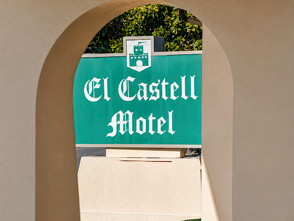 A green sign reading &ldquo;El Castell Motel&rdquo; with greenery above and a courtyard walkway below it.