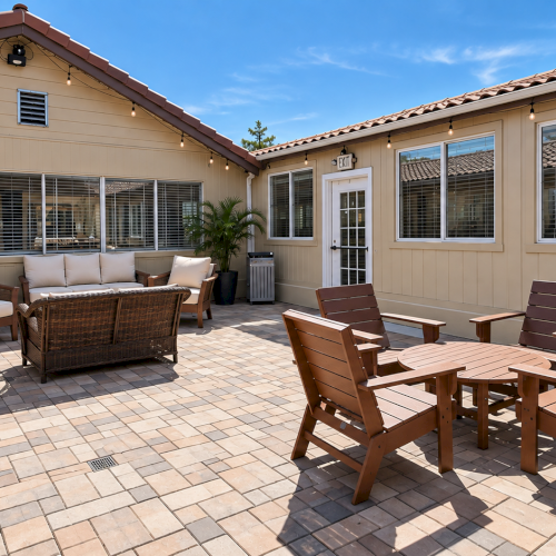 Sunlit patio with wooden chairs and table, wicker loveseat, string lights, and potted plants in front of a beige house.