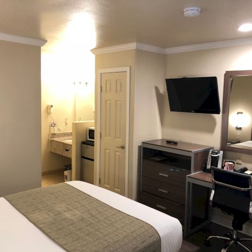 Hotel room with a bed, kitchenette counter and microwave, wall TV, and a desk with lamp, reflected in a large mirror.