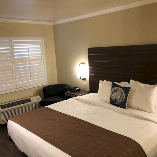 A hotel room with two beds, pillows, a bedside table and lamp, plus a window with blinds and a wall AC.