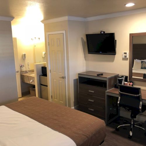 Hotel room view with a bed, desk and wall TV, plus a doorway to a small bathroom with vanity and mini-fridge.