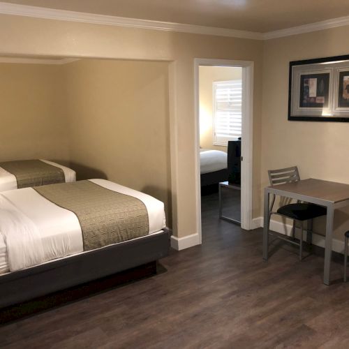 A neat hotel room with a bed, dining table and chairs, framed artwork, and an open doorway leading to another room.
