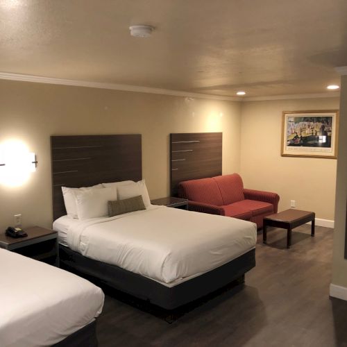 A tidy hotel room with two beds, bedside tables, wall lamps, a red upholstered chair, and framed wall art.