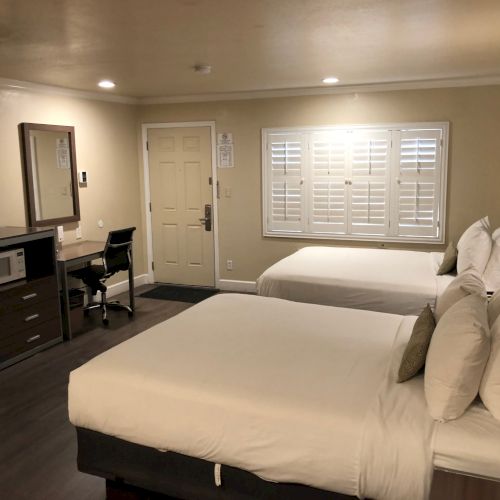 A hotel room with a bed, desk and chair, microwave/coffee station, TV, and large window with white shutters and lamps.