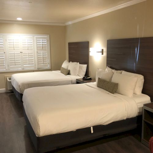 A hotel room with two twin beds, white bedding, dark headboards, nightstands, and a window with shutters.