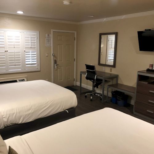 A tidy hotel room with beds, white blinds, a desk and chair, wall-mounted TV, dresser, and a door.