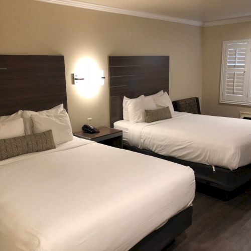 A hotel room with two neatly made beds, decorative pillows, nightstands, and a window with white blinds and curtain rods.