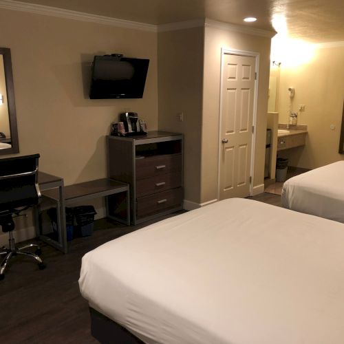 A hotel-style room with a bed, desk, chair, wall-mounted TV, coffee maker, and an open doorway to a bathroom.