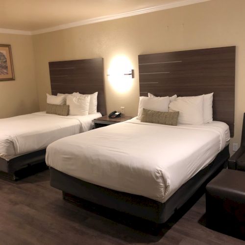 A tidy hotel room with two made-up beds, pillows, and bedside lamps against dark headboards and beige walls.