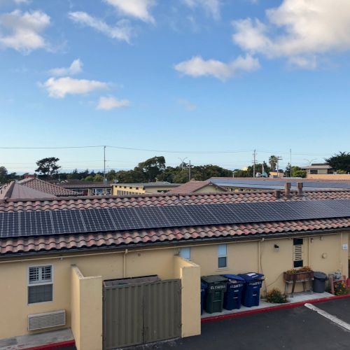 Rooftops with solar panels stretch under a bright blue sky with scattered clouds over a quiet neighborhood.