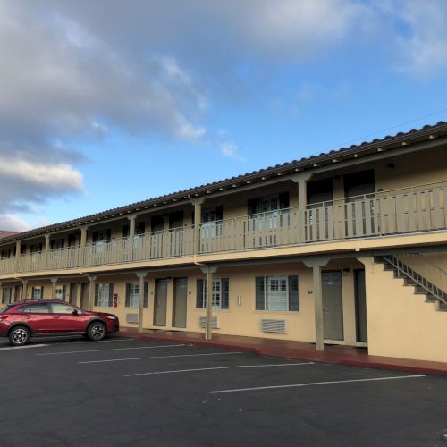 Row of beige apartment units with balconies and parking spaces under a blue sky with clouds.