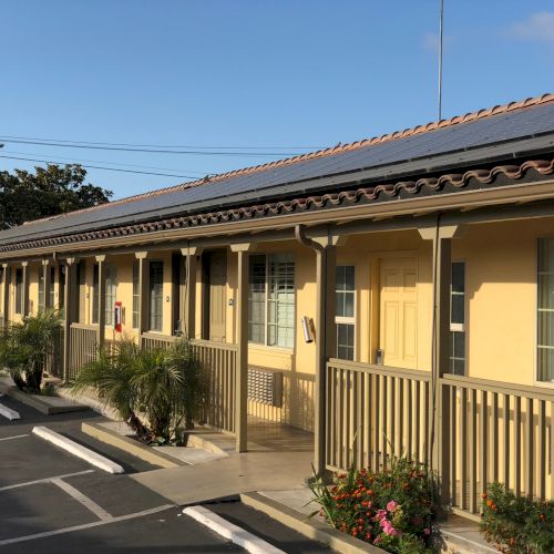 Sunlit single-story building with a row of rooftop solar panels, windows, and a sidewalk lined with plants.