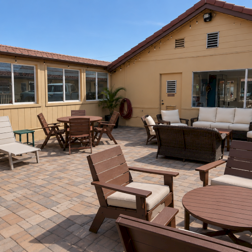 A sunny backyard patio with outdoor seating&mdash;lounge chairs, tables, and string lights&mdash;set beside sliding windows.