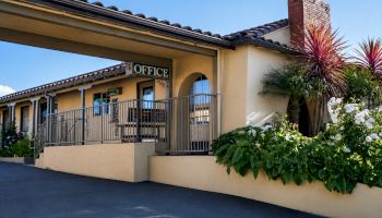 An outdoor office entrance on a sunny day, with a large &ldquo;OFFICE&rdquo; sign, arched doorway, railings, and lush plants.