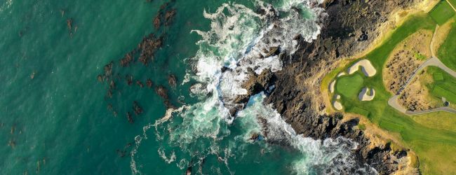 A coastal cliffside with green hills, rocky shoreline, and turquoise ocean meeting the rugged coast at the edge of land.