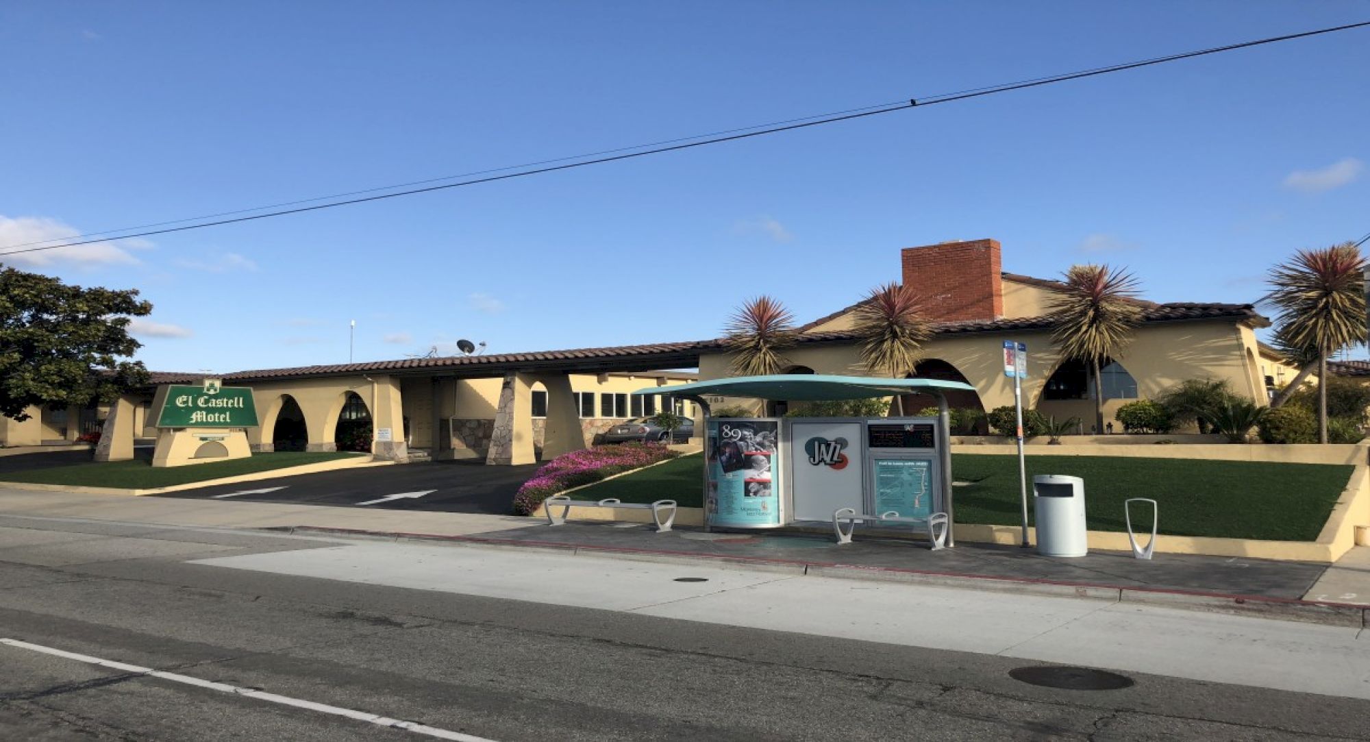 A sunny street scene with the El Cabell Hotel entrance and arches, plus a bus stop/newsstand in front.